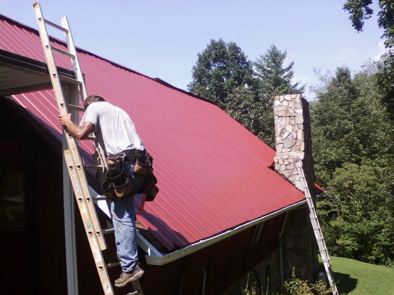 Roofer on a ladder beside a red metal roof and stone chimney — Soffit & Fascia Repair by Roofing Brothers in Bennet